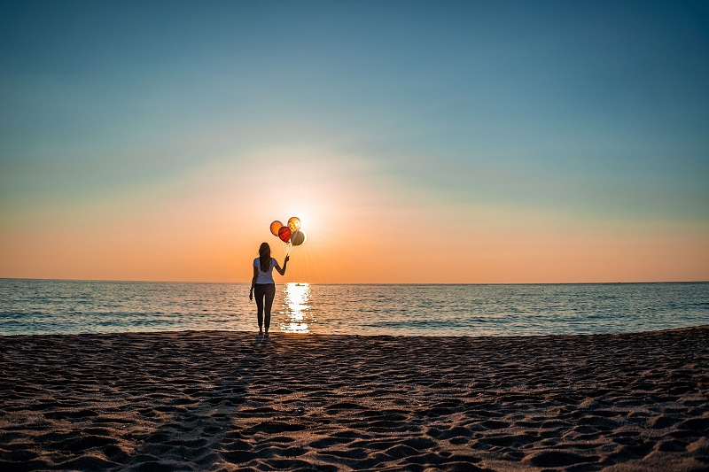 Basics of CBD Woman on a Beach Looking at the Sun Set Holding a Bunch of Balloons
