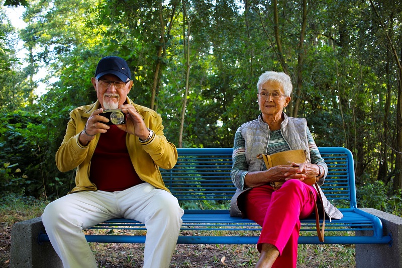 CBD for Pain Two Older People Sitting on a Bench One Holding a Camera and The Other with Crossed Arms