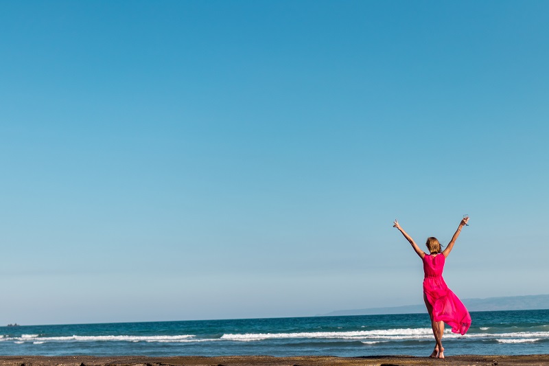 CBD Routine Woman in a Red Dress at a Beach Staring at the Ocean with Her Hands up in the Air