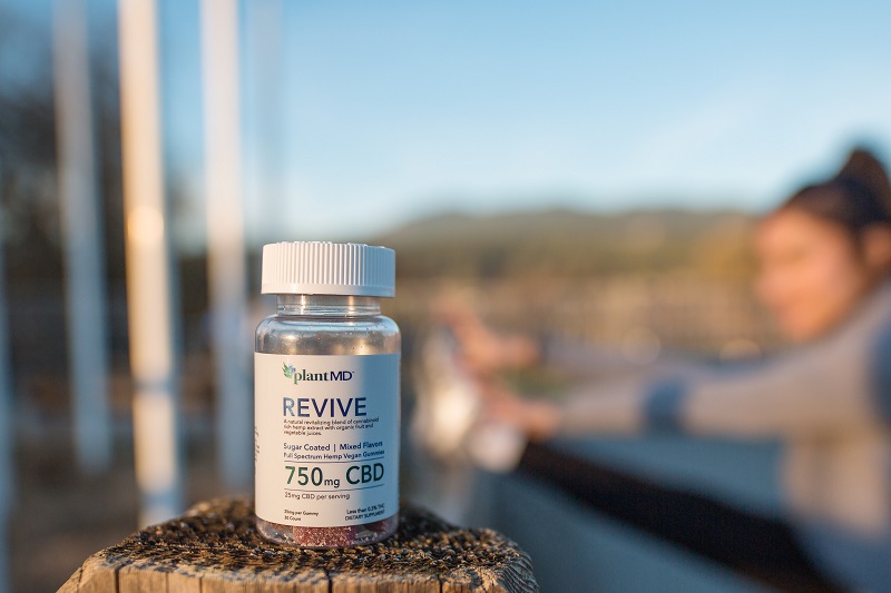 Difference Between CBD Isolate Gummies and Full Spectrum Gummies Close Up of a Bottle of PlantMD Revive Gummies on a Wodden Post Outside with a Woman in the Background Stretching