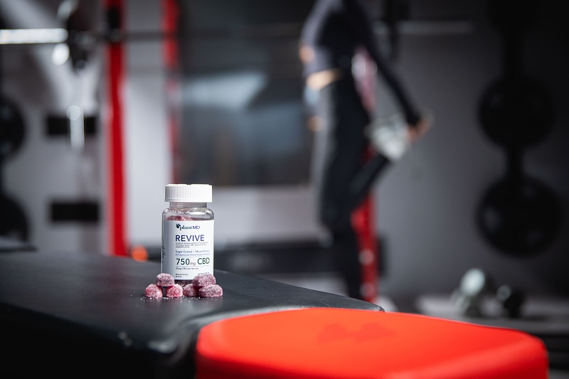 Difference Between CBD Isolate Gummies and Full Spectrum Gummies Bottle of PlantMD Revive Gummies Sitting on a Workout Bench in a Gym with a Woman Working Out in the Background