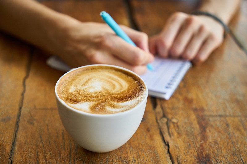 CBD Coffee Recipes a Cup of Coffee on a Table with Someone Writing on a Notepad Behind it