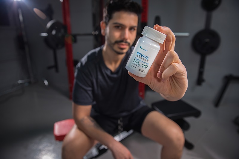 How Long Does CBD Stay in Your System a Man in a Gym Holding Up a Bottle of PlantMD Revive Capsules