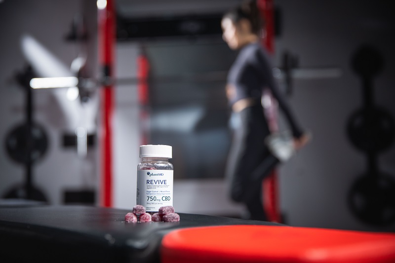 Why High-Quality Formulations Matter Most in CBD Products PlantMD Revive Gummies Sitting on a Workout Bench with a Woman Working Out in the Background